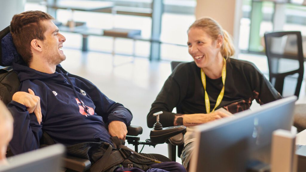 A student in a wheelchair is laughing with a support worker sat in front of a computer